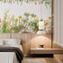 Botanical wallpaper mural featuring delicate South African flora and wildflowers in soft natural tones, with trailing greenery overhead and illustrated aloe, fynbos, and meadow plants across a light background. On a wall in a bedroom above the bed and wooden paneling. 