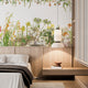 Botanical wallpaper mural featuring delicate South African flora and wildflowers in soft natural tones, with trailing greenery overhead and illustrated aloe, fynbos, and meadow plants across a light background. On a wall in a bedroom above the bed and wooden paneling. 