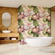 A modern bathroom interior featuring a freestanding white bathtub in front of a wall covered in a large-scale botanical print wallpaper with pink proteas and green leaves. To the left is a wood-look herringbone wall with an oval mirror and a matching wood vanity with a white sink.