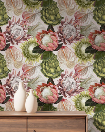 Botanical wallpaper with a seamless repeating pattern of hand painted blush pink proteas, cream anthuriums, and green foliage on a soft neutral background, styled behind a wooden cabinet with two white ceramic vases.
