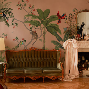 Blush pink tropical wallpaper with illustrated palms, birds, monkeys and delicate botanical details in a hand-painted chinoiserie style. On wall in a vintage living room. 