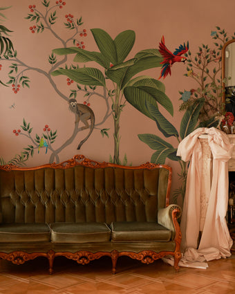 Blush pink tropical wallpaper with illustrated palms, birds, monkeys and delicate botanical details in a hand-painted chinoiserie style. On wall in a vintage living room. 