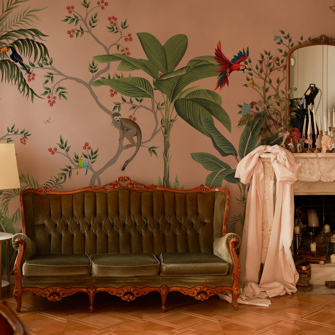 Blush pink tropical wallpaper with illustrated palms, birds, monkeys and delicate botanical details in a hand-painted chinoiserie style. On wall in a vintage living room. 