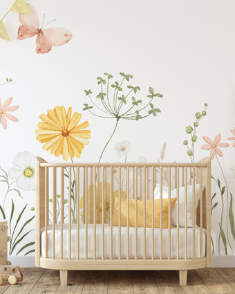 A styled nursery interior featuring a light wood crib with white bedding and yellow pillows, set against a watercolour wildflower meadow wallpaper with soft yellow and pink flowers, green foliage and delicate butterflies, and a small wooden ladder.
