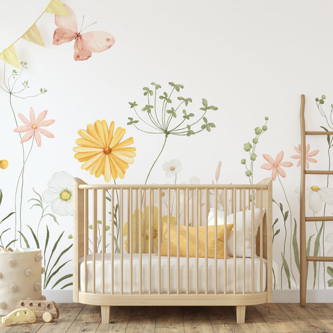A styled nursery interior featuring a light wood crib with white bedding and yellow pillows, set against a watercolour wildflower meadow wallpaper with soft yellow and pink flowers, green foliage and delicate butterflies, and a small wooden ladder.