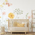 A styled nursery interior featuring a light wood crib with white bedding and yellow pillows, set against a watercolour wildflower meadow wallpaper with soft yellow and pink flowers, green foliage and delicate butterflies, and a small wooden ladder.