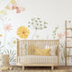 A styled nursery interior featuring a light wood crib with white bedding and yellow pillows, set against a watercolour wildflower meadow wallpaper with soft yellow and pink flowers, green foliage and delicate butterflies, and a small wooden ladder.