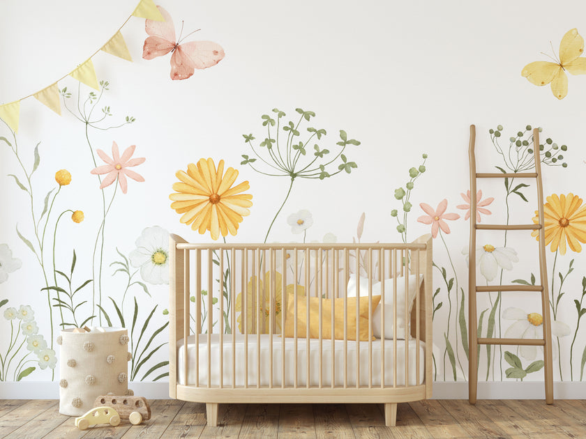 A styled nursery interior featuring a light wood crib with white bedding and yellow pillows, set against a watercolour wildflower meadow wallpaper with soft yellow and pink flowers, green foliage and delicate butterflies, and a small wooden ladder.