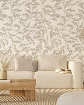 A styled living room scene featuring a cream coloured sofa against a wall with white botanical wallpaper featuring finely illustrated taupe camellia branches and delicate leaves. There is a wooden coffee table, a floor lamp, and a light rug.

