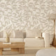 A styled living room scene featuring a cream coloured sofa against a wall with white botanical wallpaper featuring finely illustrated taupe camellia branches and delicate leaves. There is a wooden coffee table, a floor lamp, and a light rug.

