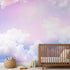 Realistic nursery scene with a playful sky mural in pinks, purples, and blues covering the entire wall, illuminated by soft daylight.