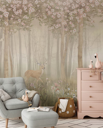 Children's woodland mural featuring a deer in a misty forest with wildflowers and flowering trees in soft blush and neutral tones, creating a dreamy nursery setting. In the room there is a light grey armchair, pink dresser, and other decorative items.
