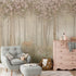 Children's woodland mural featuring a deer in a misty forest with wildflowers and flowering trees in soft blush and neutral tones, creating a dreamy nursery setting. In the room there is a light grey armchair, pink dresser, and other decorative items.
