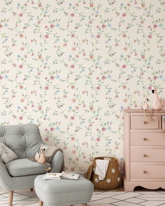 A styled nursery room featuring delicate pink and blue floral wallpaper with trailing green stems and leaves on a warm cream background. The room is furnished with a light grey tufted armchair and matching ottoman, a dusty pink wooden dresser, and a woven basket.