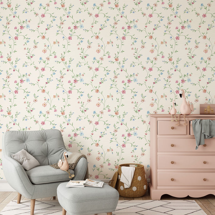 A styled nursery room featuring delicate pink and blue floral wallpaper with trailing green stems and leaves on a warm cream background. The room is furnished with a light grey tufted armchair and matching ottoman, a dusty pink wooden dresser, and a woven basket.