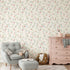 A styled nursery room featuring delicate pink and blue floral wallpaper with trailing green stems and leaves on a warm cream background. The room is furnished with a light grey tufted armchair and matching ottoman, a dusty pink wooden dresser, and a woven basket.