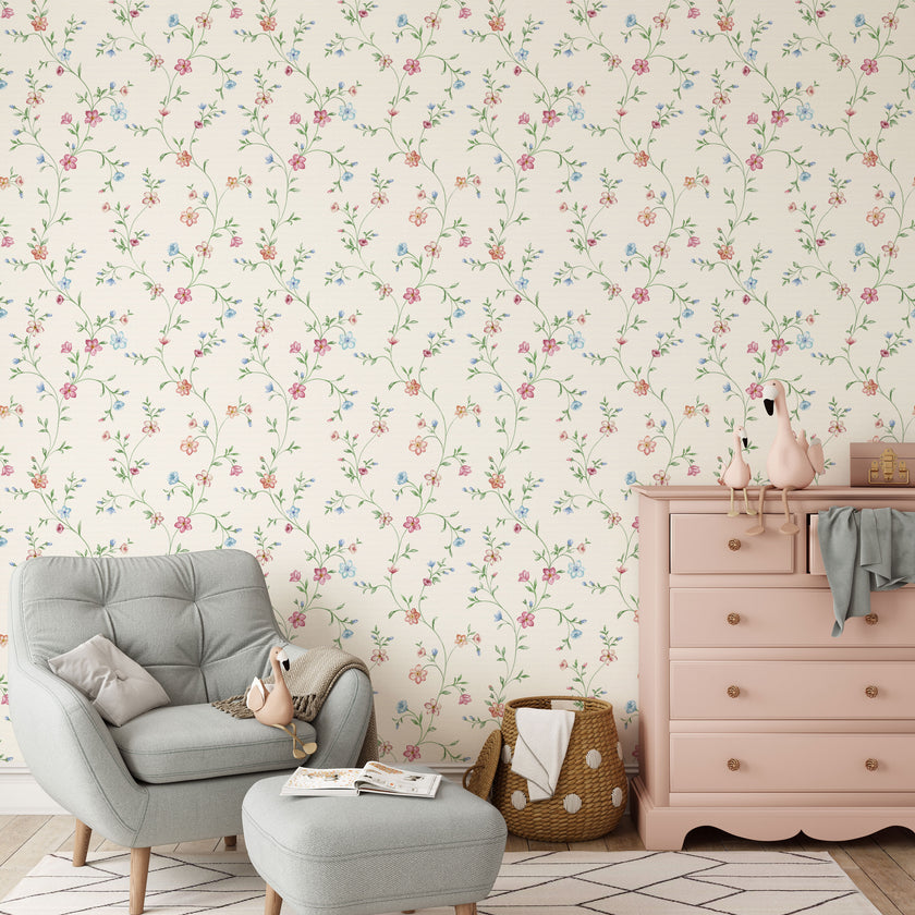 A styled nursery room featuring delicate pink and blue floral wallpaper with trailing green stems and leaves on a warm cream background. The room is furnished with a light grey tufted armchair and matching ottoman, a dusty pink wooden dresser, and a woven basket.