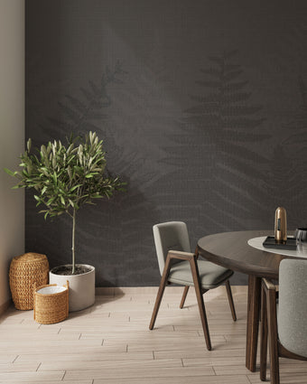 A styled interior corner featuring a charcoal grey botanical mural featuring oversized layered fern fronds on a textured grey background. With light wood flooring, a potted olive tree, woven baskets, and a round wooden dining table with grey upholstered chairs.
