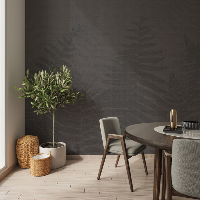 A styled interior corner featuring a charcoal grey botanical mural featuring oversized layered fern fronds on a textured grey background. With light wood flooring, a potted olive tree, woven baskets, and a round wooden dining table with grey upholstered chairs.