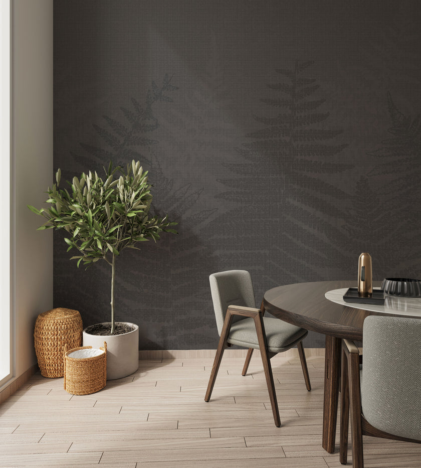 A styled interior corner featuring a charcoal grey botanical mural featuring oversized layered fern fronds on a textured grey background. With light wood flooring, a potted olive tree, woven baskets, and a round wooden dining table with grey upholstered chairs.