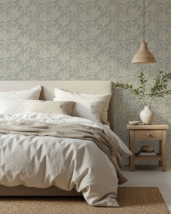 A styled bedroom scene featuring a bed dressed in neutral beige and white linens, set against a wall with a sage grey botanical wallpaper with a seamless repeat, featuring ivory stylised floral blooms and delicate foliage with hand drawn detailing.