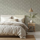 A styled bedroom scene featuring a bed dressed in neutral beige and white linens, set against a wall with a sage grey botanical wallpaper with a seamless repeat, featuring ivory stylised floral blooms and delicate foliage with hand drawn detailing.