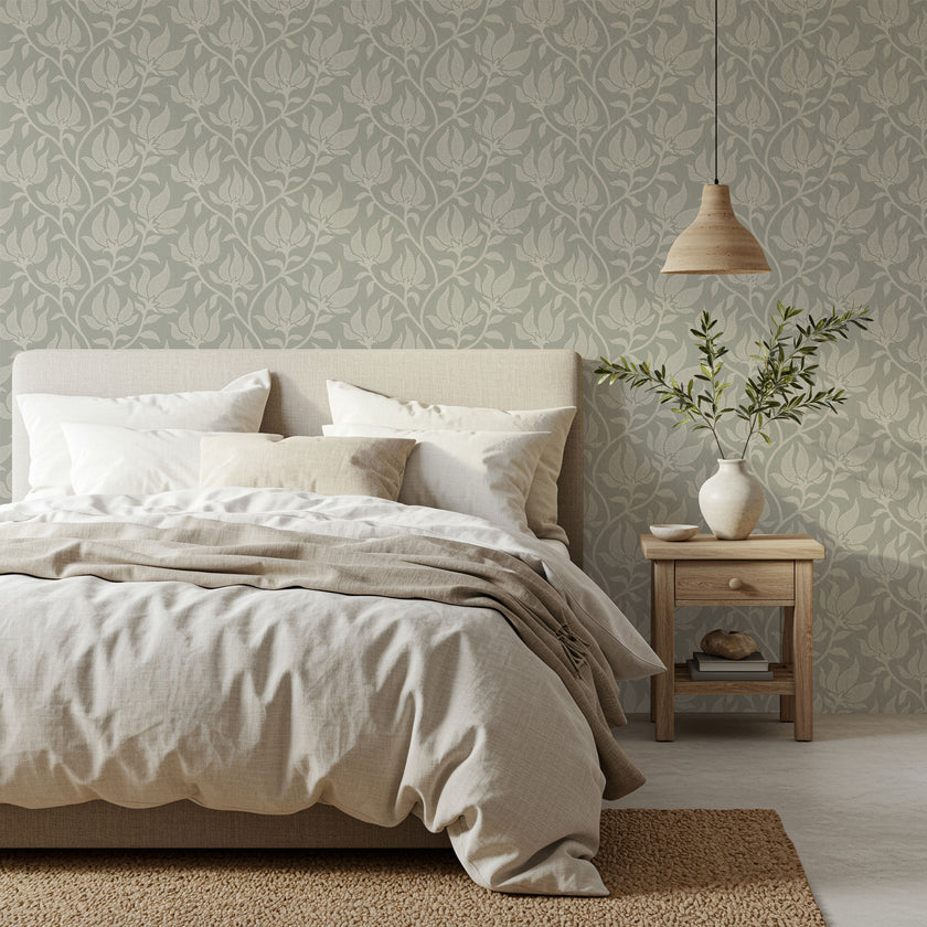 A styled bedroom scene featuring a bed dressed in neutral beige and white linens, set against a wall with a sage grey botanical wallpaper with a seamless repeat, featuring ivory stylised floral blooms and delicate foliage with hand drawn detailing.