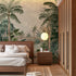 A styled bedroom interior featuring a tropical mural with lush green palms and foliage surrounding a blue lagoon with soft mountains in the background, behind a bed. The room includes a wooden bed frame, white bedding, a matching wooden nightstand with a round white pendant light, a slatted wood wardrobe, and light wood flooring.

