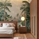 A styled bedroom interior featuring a tropical mural with lush green palms and foliage surrounding a blue lagoon with soft mountains in the background, behind a bed. The room includes a wooden bed frame, white bedding, a matching wooden nightstand with a round white pendant light, a slatted wood wardrobe, and light wood flooring.

