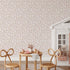A styled interior scene featuring a
soft pink and neutral floral wallpaper with delicate flowers and leafy sprigs on a subtle striped background., with a small wooden table, two woven chairs, a white playhouse, and pink rain boots on the floor.