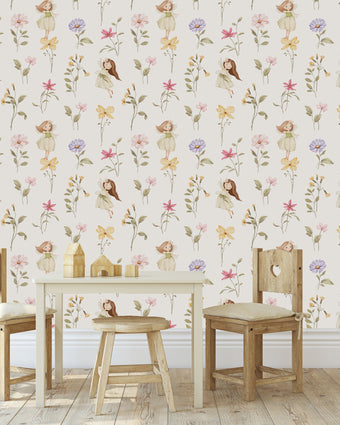 A styled children's playroom featuring a Whimsical little fairy wallpaper with delicate pink, lilac and yellow wildflowers and soft green foliage. In front of the wall is a small wooden table, two wooden chairs with cream cushions, and a small stool.
