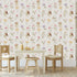 A styled children's playroom featuring a Whimsical little fairy wallpaper with delicate pink, lilac and yellow wildflowers and soft green foliage. In front of the wall is a small wooden table, two wooden chairs with cream cushions, and a small stool.