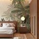 A styled bedroom interior featuring a tropical mural with layered palm trees and lush green foliage on a soft neutral background on the back wall, a wooden bed with white bedding, a matching wooden nightstand and wardrobe, and a round white pendant light.

