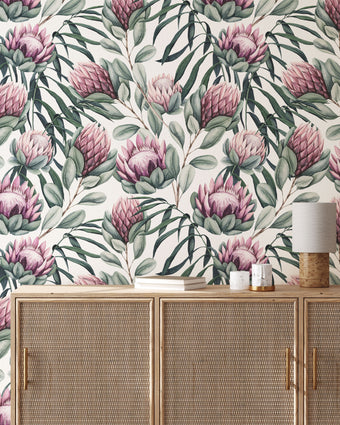 Botanical wallpaper with a seamless repeating pattern of hand painted king proteas, eucalyptus leaves, and soft sage green foliage on a white background. Shown above a light wood cabinet with woven doors and a small table lamp.