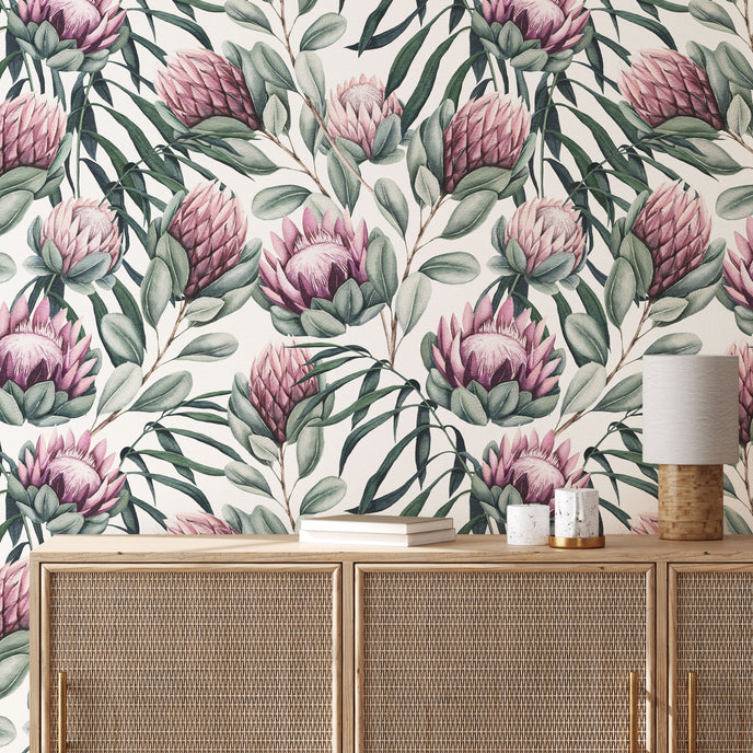 Botanical wallpaper with a seamless repeating pattern of hand painted king proteas, eucalyptus leaves, and soft sage green foliage on a white background. Shown above a light wood cabinet with woven doors and a small table lamp.