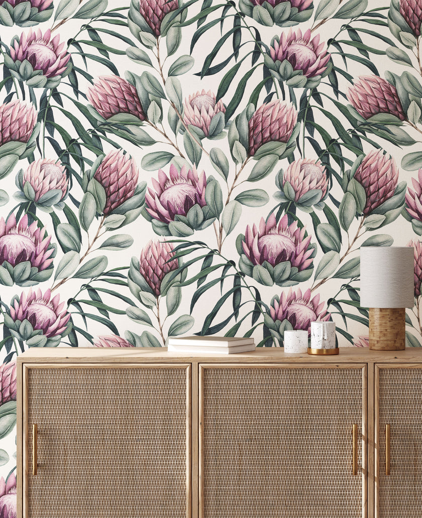 Botanical wallpaper with a seamless repeating pattern of hand painted king proteas, eucalyptus leaves, and soft sage green foliage on a white background. Shown above a light wood cabinet with woven doors and a small table lamp.