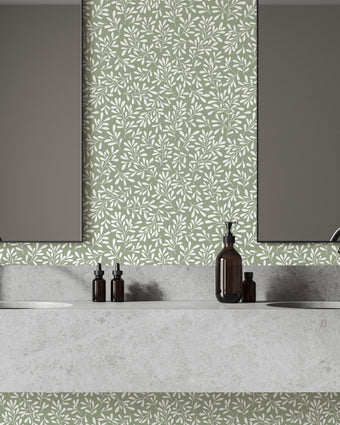 A modern bathroom vanity setup featuring a light grey stone double sink countertop with two dark faucets, two rectangular mirrors, and a sage green botanical wallpaper with a seamless repeat, featuring white leafy sprigs in a simple, elegant pattern.
