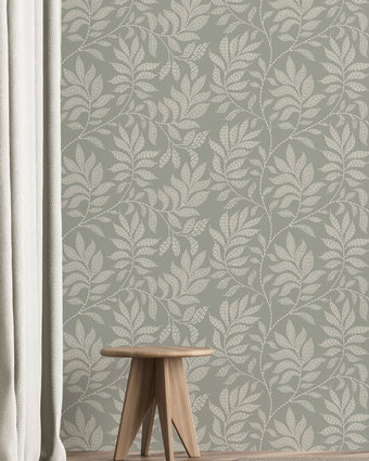 A styled interior scene showing a wall covered in a repeating botanical leaf pattern in muted sage green and off-white. A small wooden stool sits on a light wood floor, with a white curtain on the left and part of a rug in the foreground.