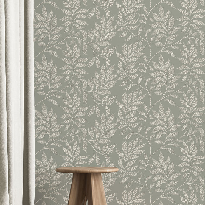 A styled interior scene showing a wall covered in a repeating botanical leaf pattern in muted sage green and off-white. A small wooden stool sits on a light wood floor, with a white curtain on the left and part of a rug in the foreground.