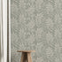 A styled interior scene showing a wall covered in a repeating botanical leaf pattern in muted sage green and off-white. A small wooden stool sits on a light wood floor, with a white curtain on the left and part of a rug in the foreground.
