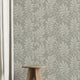 A styled interior scene showing a wall covered in a repeating botanical leaf pattern in muted sage green and off-white. A small wooden stool sits on a light wood floor, with a white curtain on the left and part of a rug in the foreground.