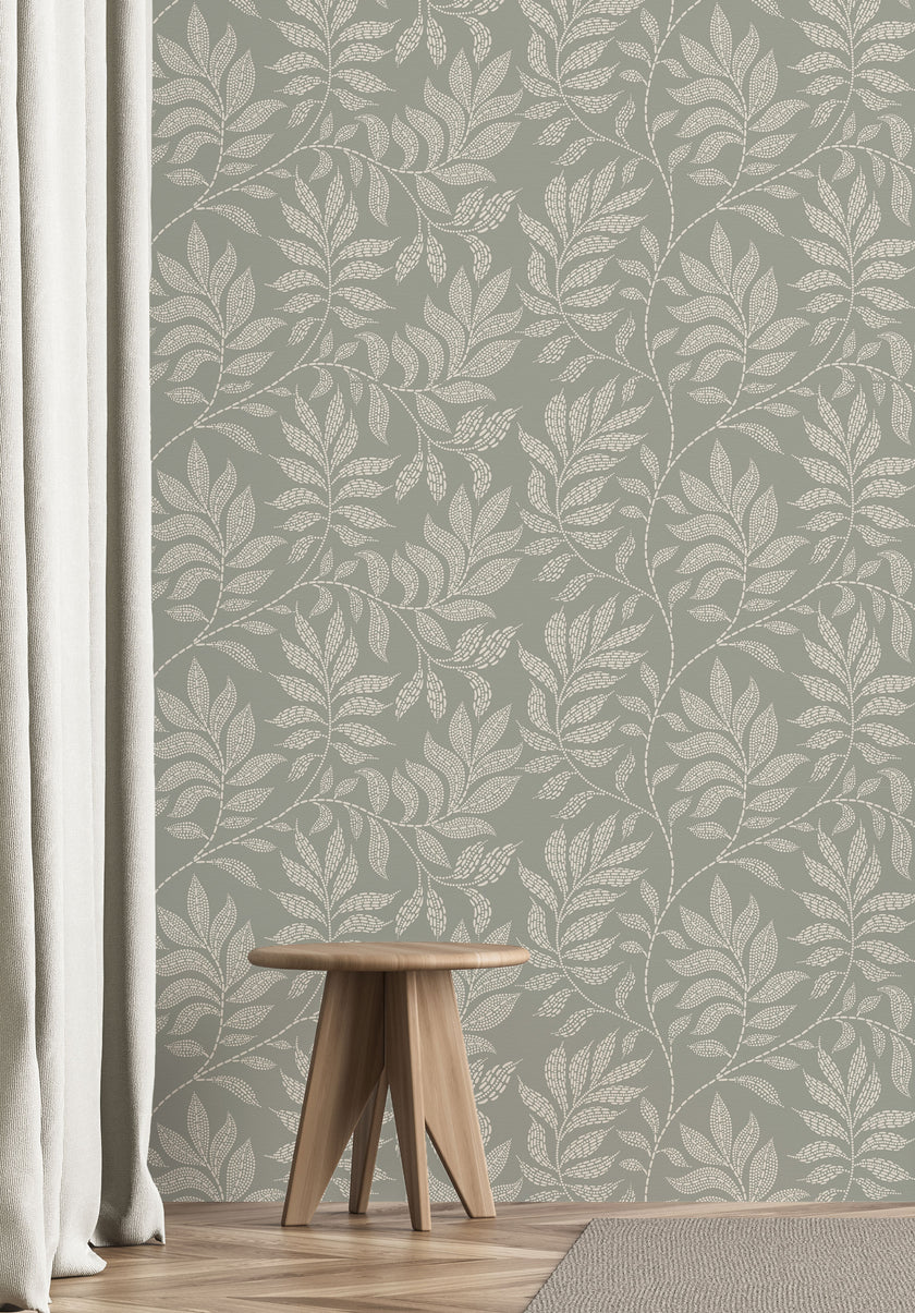 A styled interior scene showing a wall covered in a repeating botanical leaf pattern in muted sage green and off-white. A small wooden stool sits on a light wood floor, with a white curtain on the left and part of a rug in the foreground.