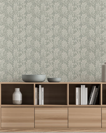 A styled interior scene showing a light wood media console against a wall with a sage grey botanical wallpaper with a seamless repeat, featuring ivory hand-drawn leaves and flowing stems with delicate dotted detailing. Decorative bowls, books, and vases sit on and inside the console.

