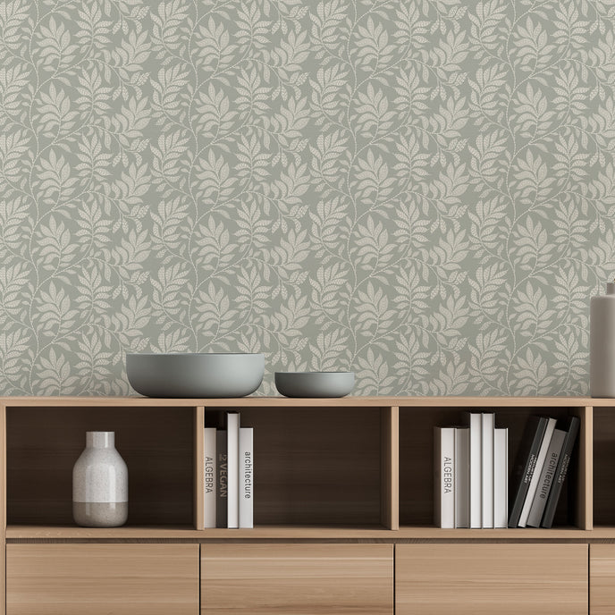 A styled interior scene showing a light wood media console against a wall with a sage grey botanical wallpaper with a seamless repeat, featuring ivory hand-drawn leaves and flowing stems with delicate dotted detailing. Decorative bowls, books, and vases sit on and inside the console.

