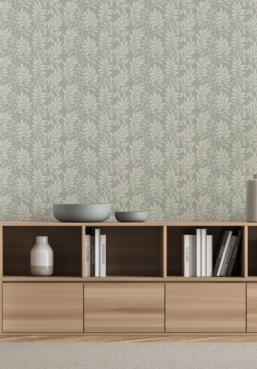 A styled interior scene showing a light wood media console against a wall with a sage grey botanical wallpaper with a seamless repeat, featuring ivory hand-drawn leaves and flowing stems with delicate dotted detailing. Decorative bowls, books, and vases sit on and inside the console.

