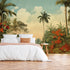 Vintage tropical mural with palm trees, lush green foliage and red orange flowers beneath a soft blue sky, displayed on the wall behind a neatly made bed with white linens and a orange throw blanket, set in a bedroom with wooden flooring.