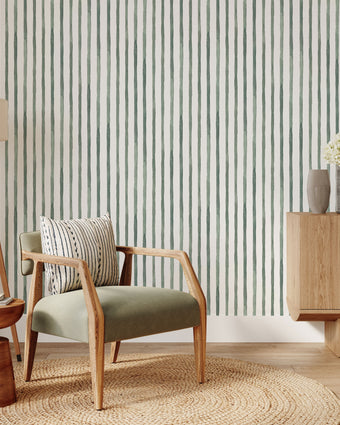 A styled interior room scene featuring  watercolour hand painted vertical stripes in sage green with a white background. In the foreground, there is a light wood armchair with a sage green upholstered seat, a striped throw pillow, a round woven jute rug, a wooden tripod floor lamp, and a matching wooden sideboard with a vase of flowers.
