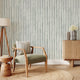 A styled interior room scene featuring  watercolour hand painted vertical stripes in sage green with a white background. In the foreground, there is a light wood armchair with a sage green upholstered seat, a striped throw pillow, a round woven jute rug, a wooden tripod floor lamp, and a matching wooden sideboard with a vase of flowers.
