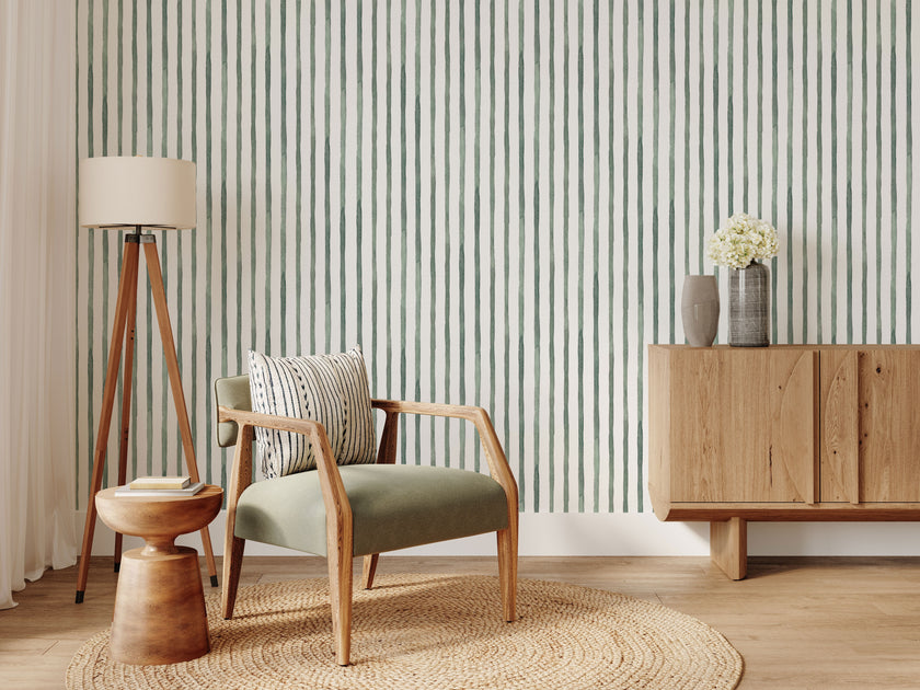 A styled interior room scene featuring  watercolour hand painted vertical stripes in sage green with a white background. In the foreground, there is a light wood armchair with a sage green upholstered seat, a striped throw pillow, a round woven jute rug, a wooden tripod floor lamp, and a matching wooden sideboard with a vase of flowers.
