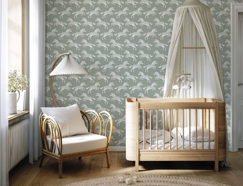 A styled nursery interior with a soft sage green wallpaper featuring a repeating pattern of detailed white zebras. With a light wood crib with white bedding and a white canopy, an armchair with a striped pillow, and a floor lamp near a window.